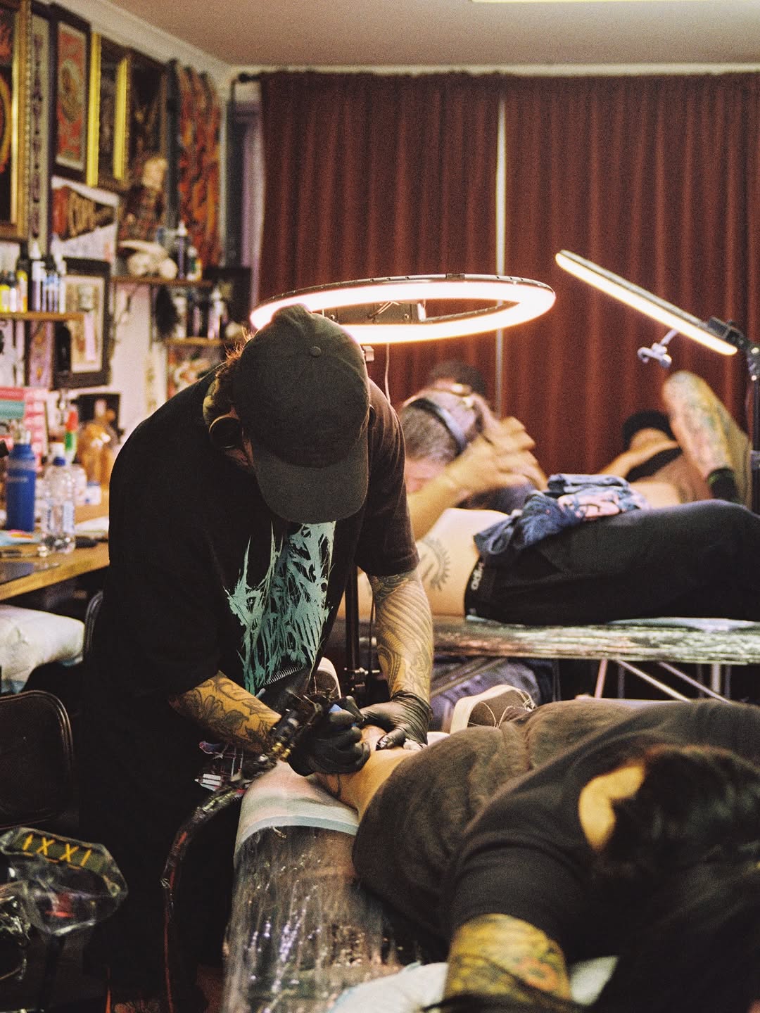 Tattoo artist working on a client's leg with a neon ring light overhead in a studio decorated with posters and artwork.