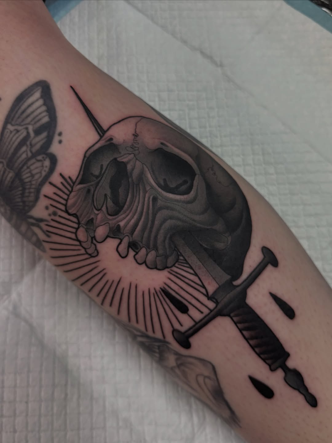 Skull with butterfly and dagger tattoo on arm, featuring detailed shading and line work in black ink.