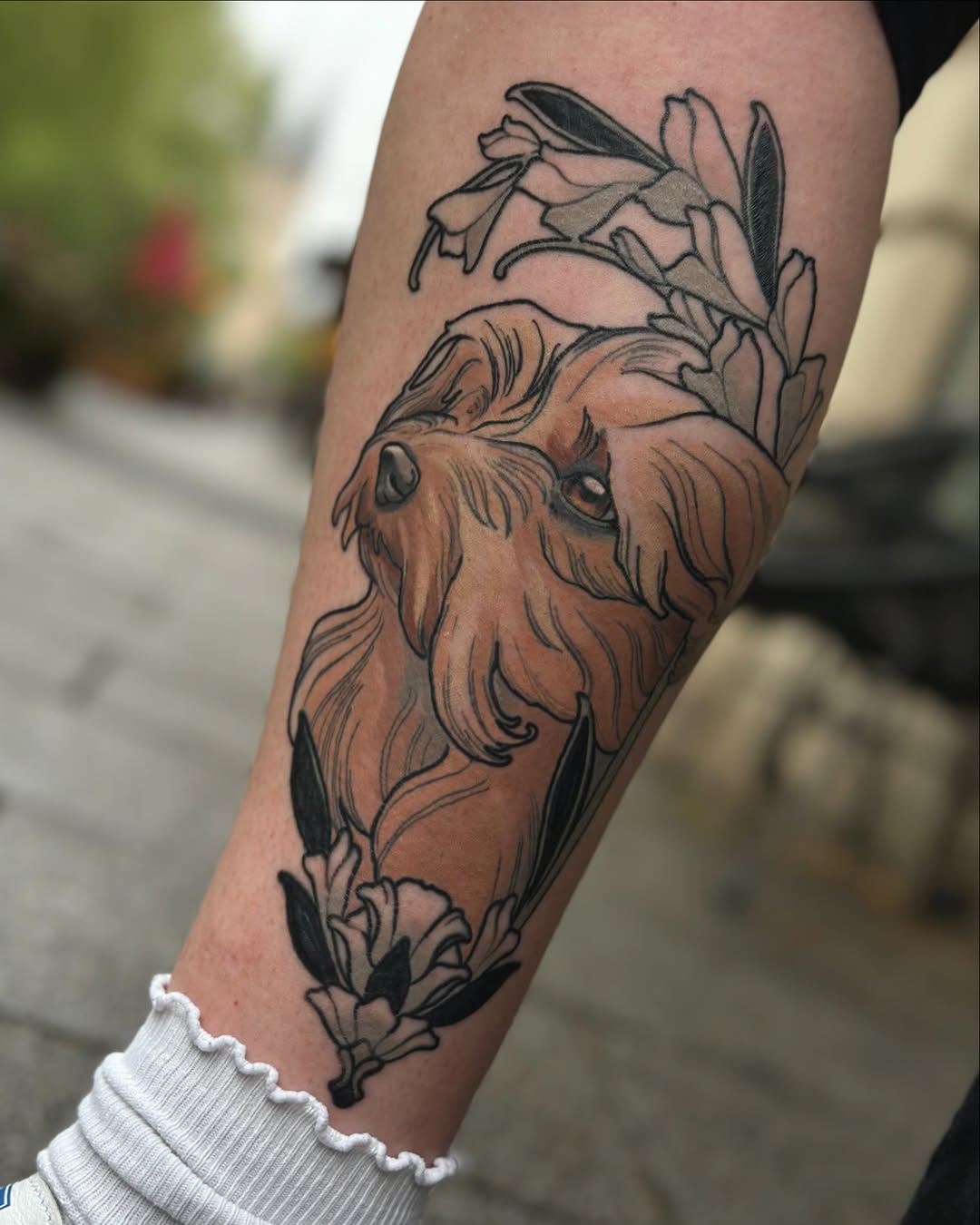 Goat head with flowing beard surrounded by flowering plants and leaves in brown and black ink on the forearm.