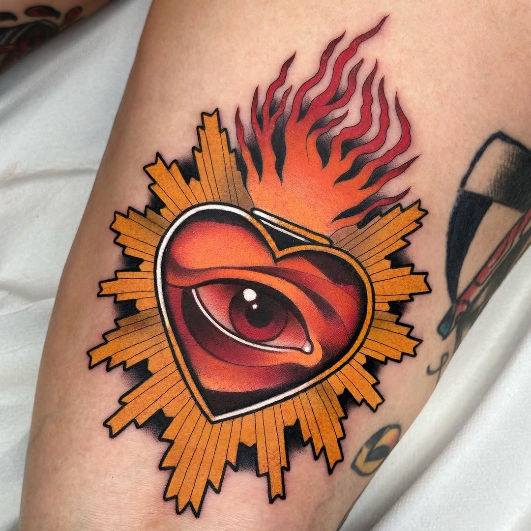 Red and orange flaming heart with an eye inside, surrounded by a gold sunburst shape, tattooed on an arm.
