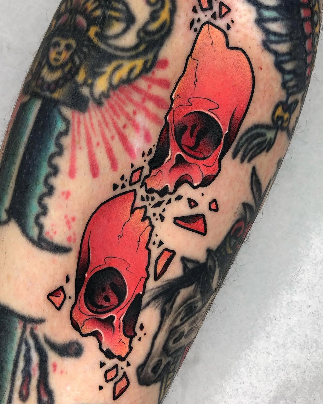 Two red cracked skulls with black outlines and scattered fragments on an upper arm, surrounded by other tattoos in darker colors.