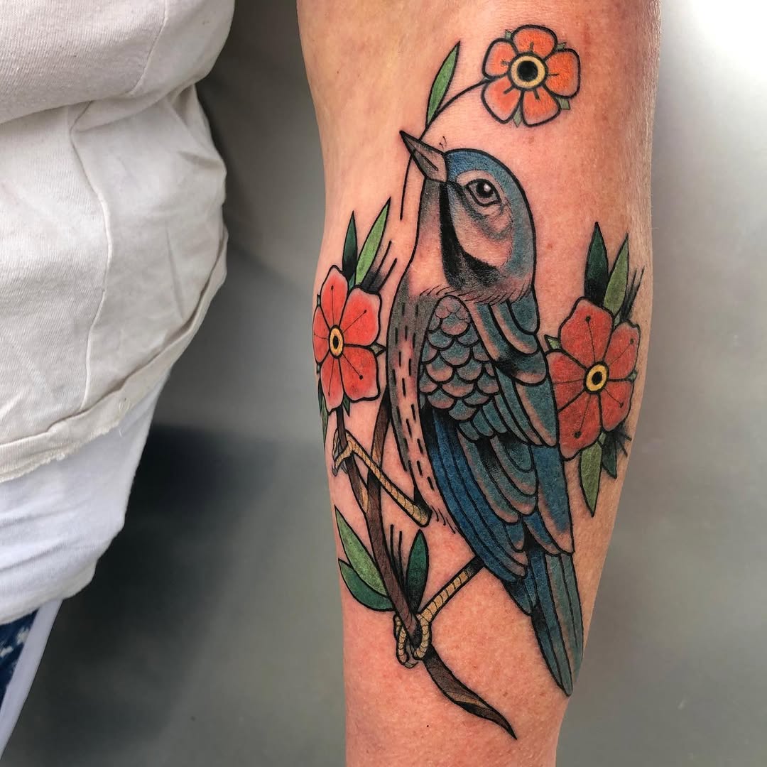 Blue bird perched on a branch with red and orange flowers and green leaves surrounding it on the forearm.