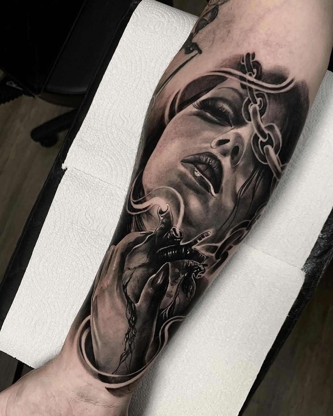 Detailed monochrome arm tattoo featuring a woman's face with ornamental chains and a heart with thorns, showcasing shading and portraiture work.