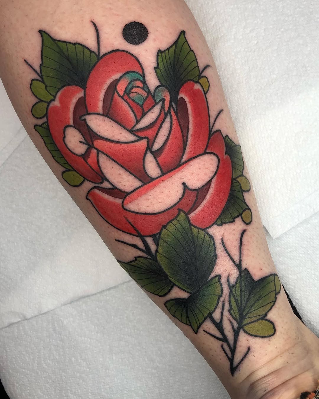 Red rose with layered petals and light pink highlights, surrounded by green leaves on a stem, tattooed on the forearm with a small black dot above.