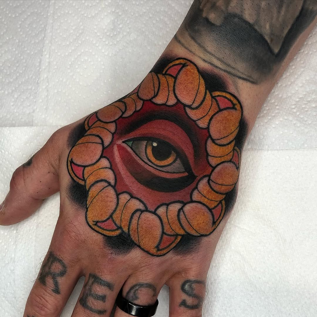 Colorful eye tattoo with orange and red tones surrounded by circular ornamental elements on the back of a hand.