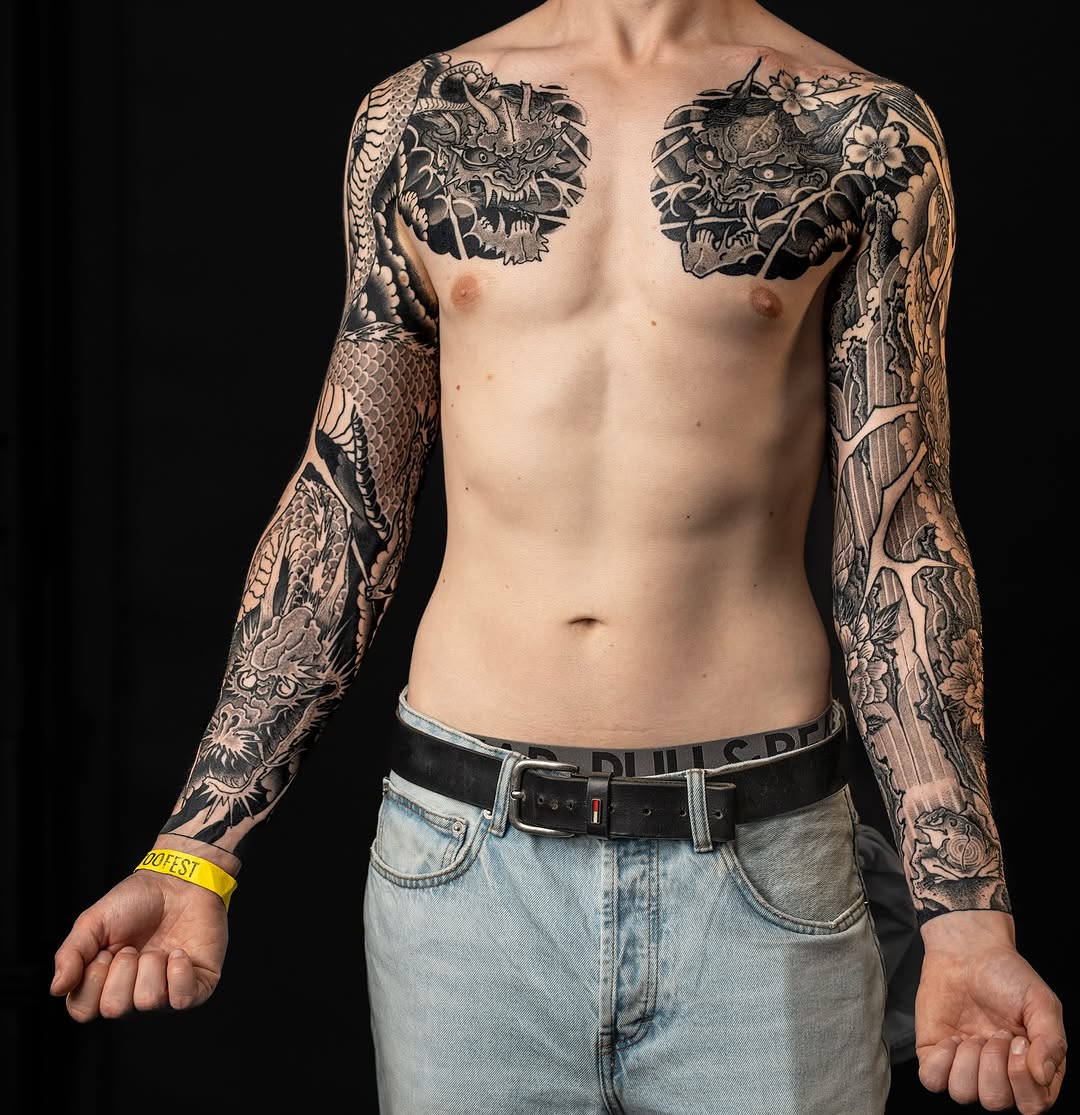 Shirtless man displaying full black sleeve tattoos on both arms featuring intricate demon and serpent designs with detailed floral elements on chest and forearms.