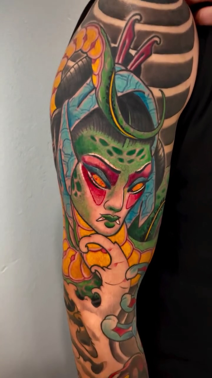 Colorful tattoo on arm featuring a green-faced figure with red markings, orange and yellow florals, and purple tentacle-like appendages extending upward.