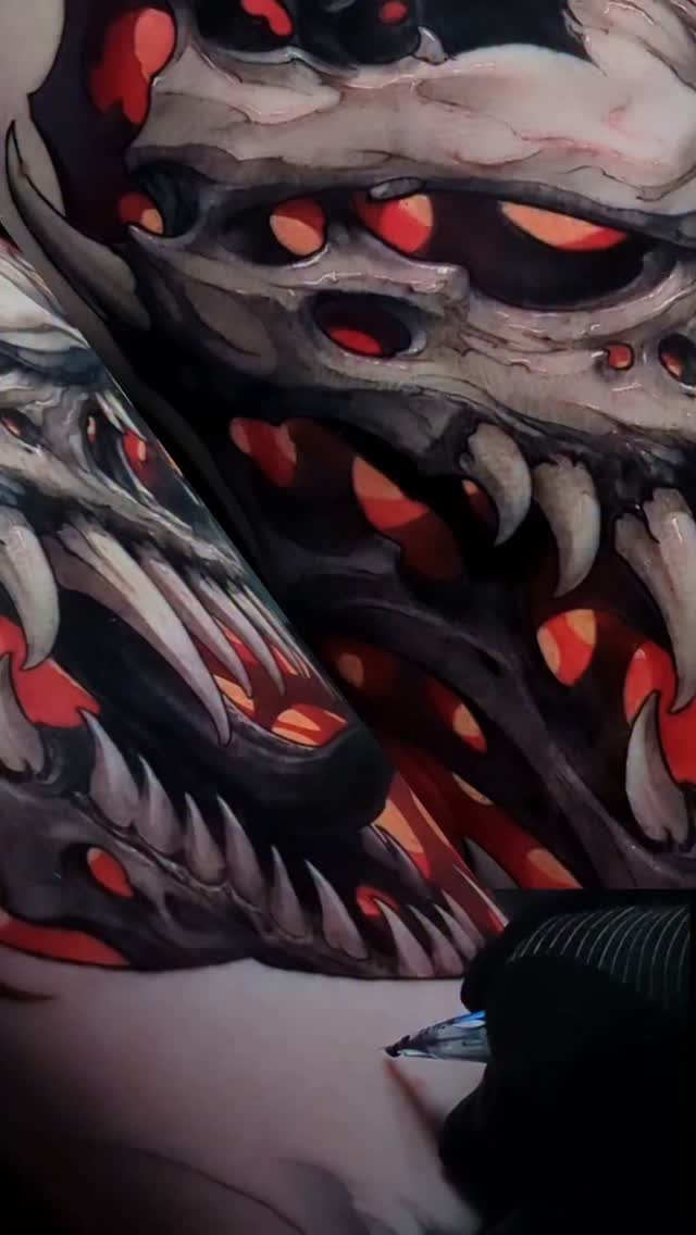 Close-up of a demon or creature face tattoo featuring dark gray and black shading with glowing red and orange eyes on an arm.