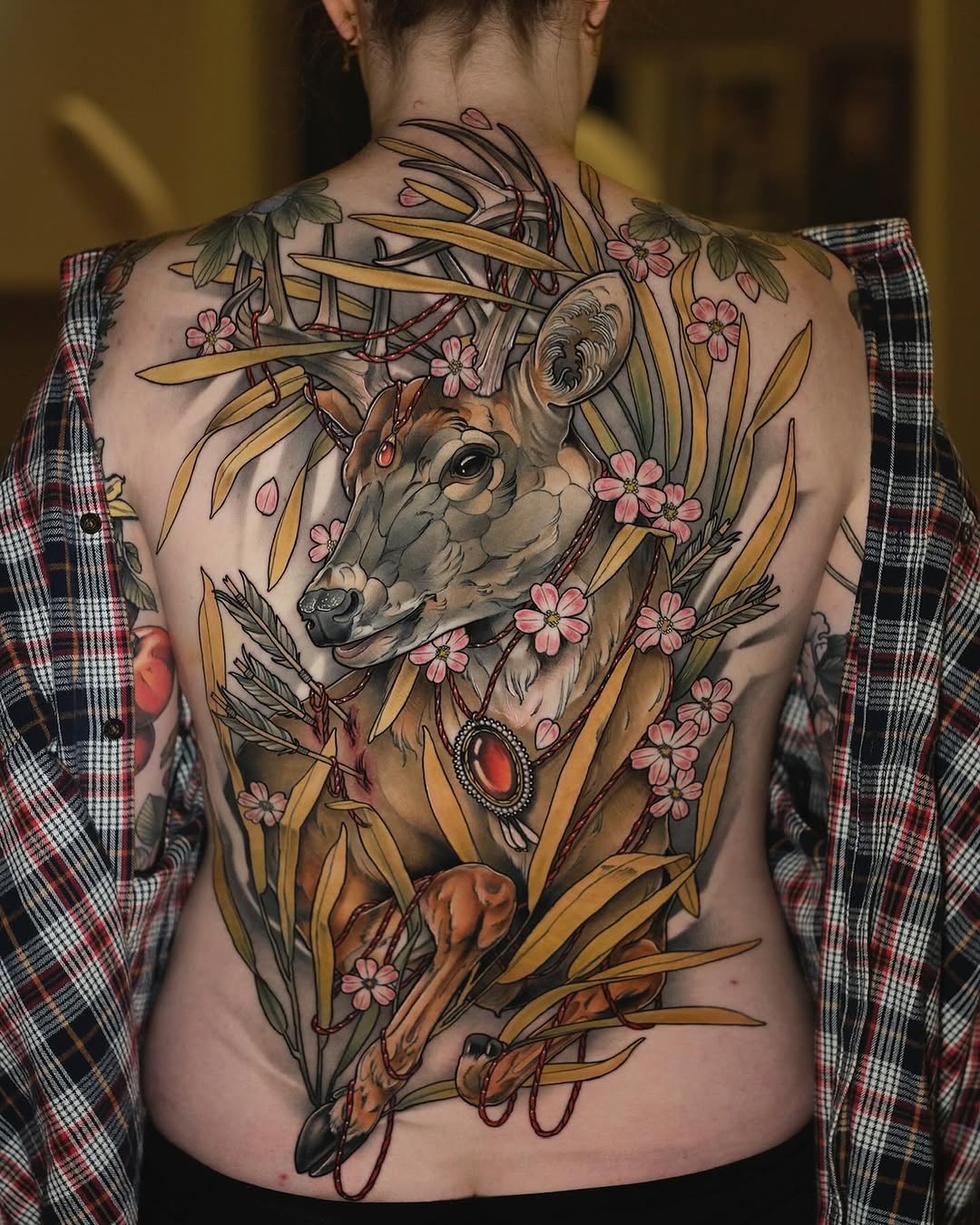 Back tattoo featuring a deer head with antlers surrounded by golden wheat stalks, pink flowers, and red gemstone accents in warm earth tones.
