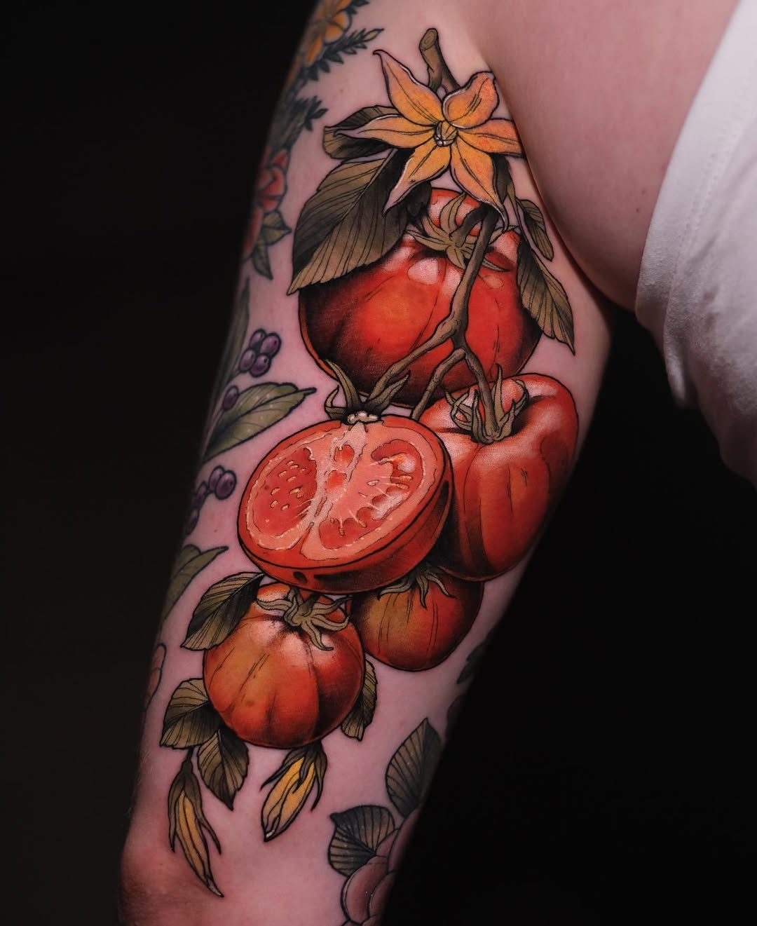 Colorful arm tattoo featuring red tomatoes at various stages with yellow flowers and green leaves against pink skin.