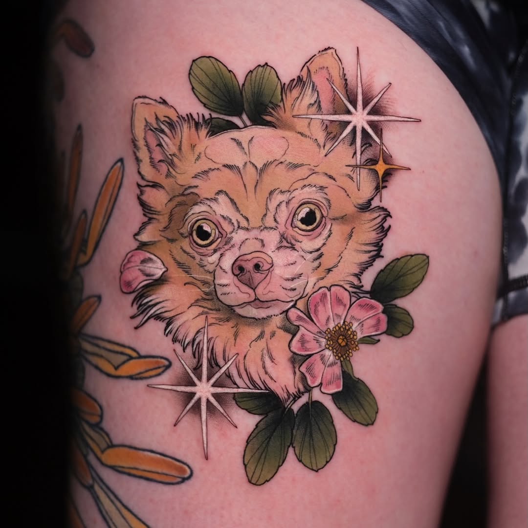 Colorful shoulder tattoo of a wide-eyed chihuahua surrounded by pink flowers, green leaves, and geometric star shapes in orange, tan, and pink tones.