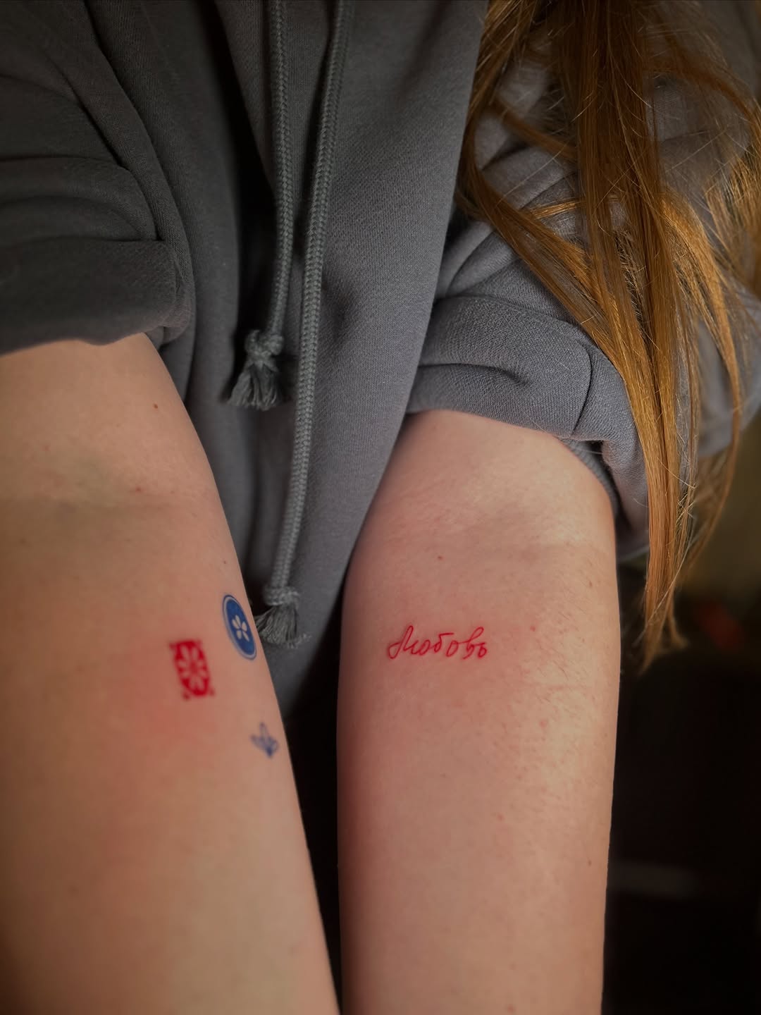 Two matching red and blue tattoos on the inner arms of a person wearing a gray hoodie, featuring geometric and text designs.