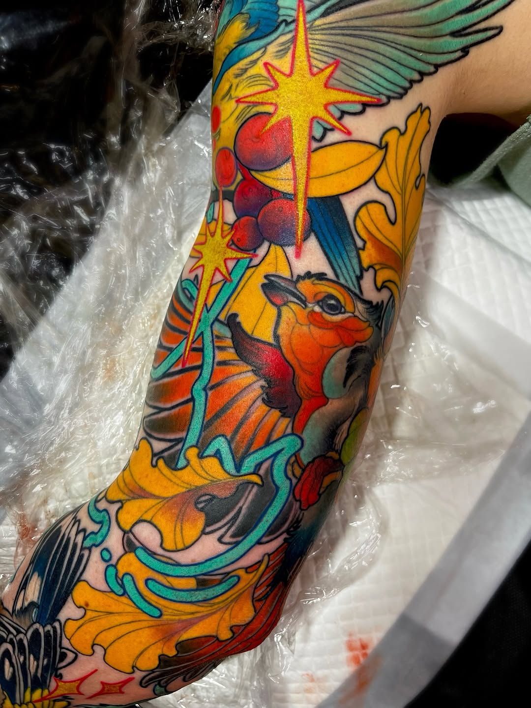 Colorful full-length sleeve tattoo featuring winged creatures, stars, ornamental scrollwork, and botanical elements in yellow, red, blue, and orange on the arm.