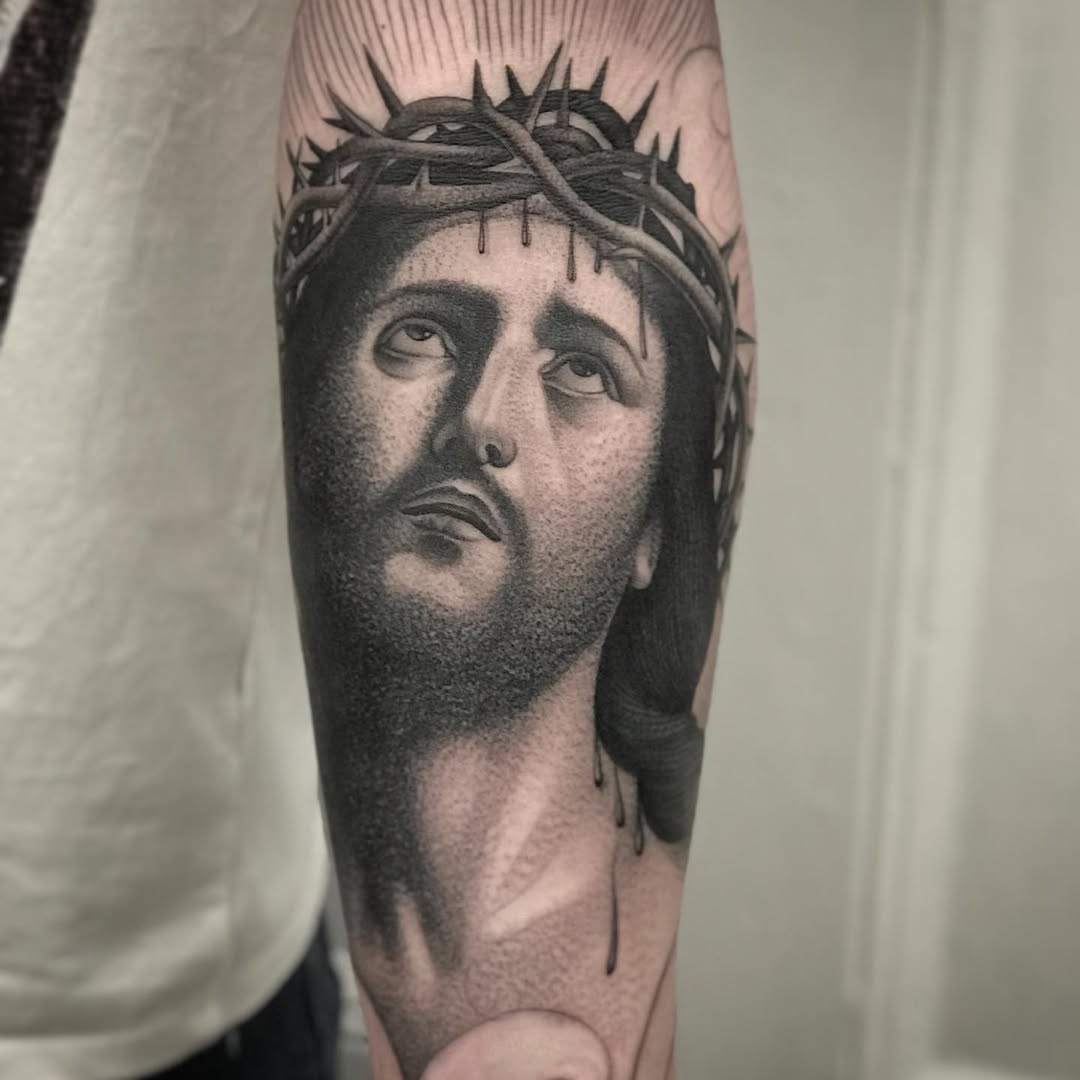 Monochromatic portrait tattoo on forearm depicting a bearded man's face wearing a crown of thorns with dripping details.