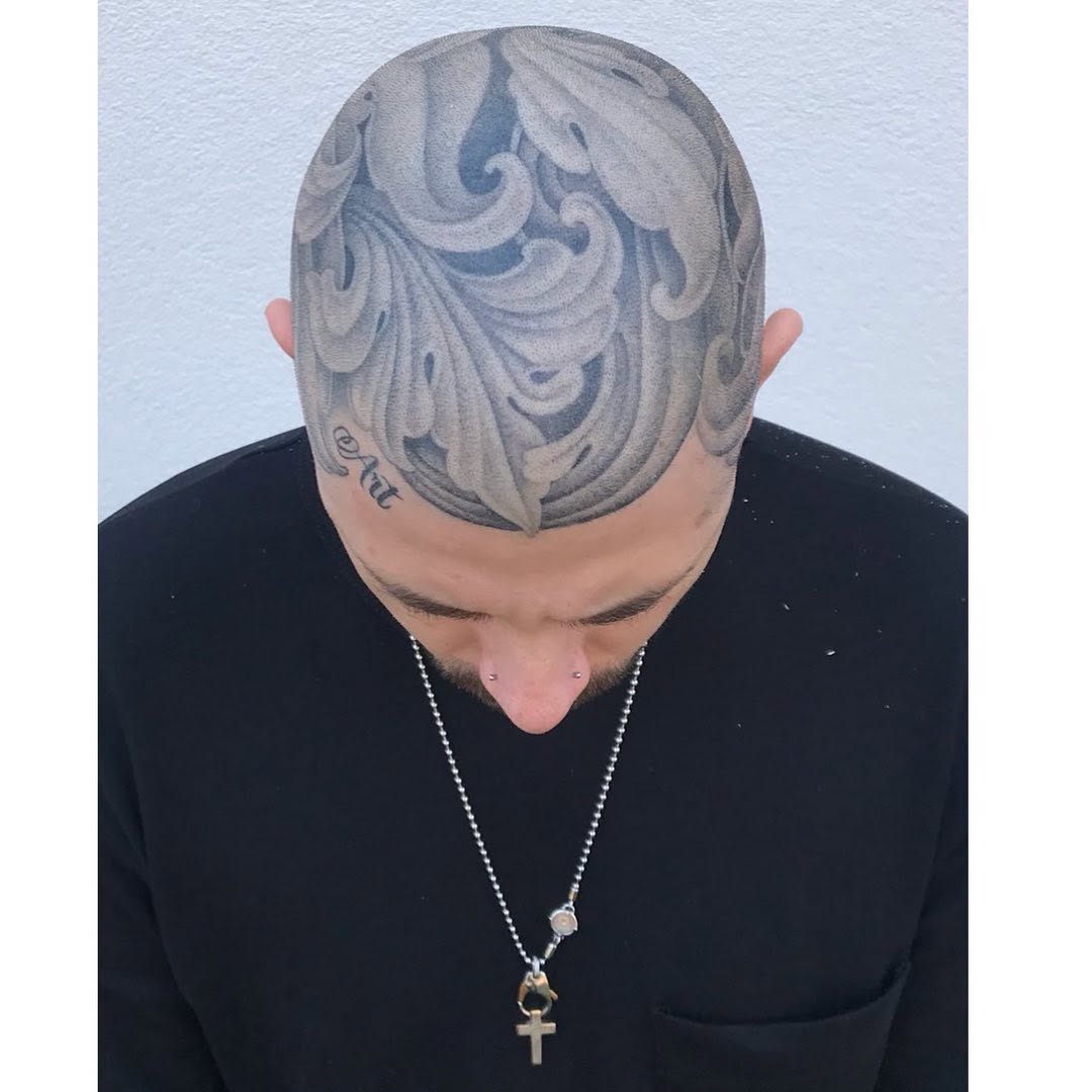 Back of head tattoo featuring ornate scrolling patterns and draped fabric designs in gray and black ink.