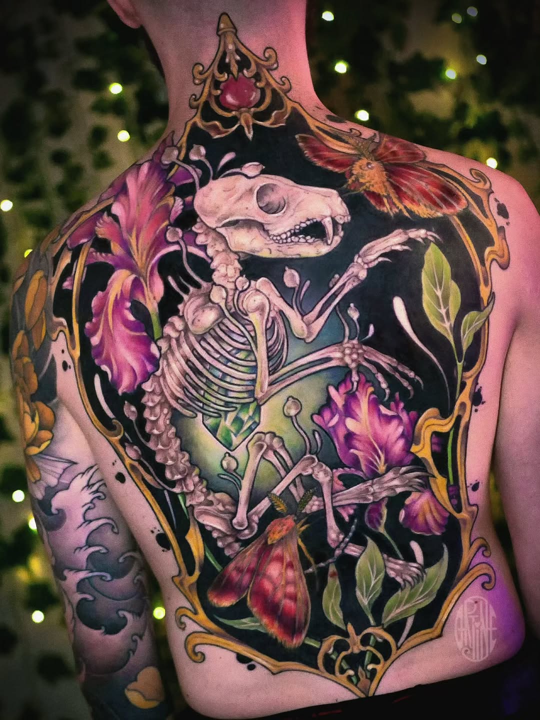 Skeletal figure surrounded by vibrant pink and orange flowers with ornamental gold borders tattooed across the back and shoulders.