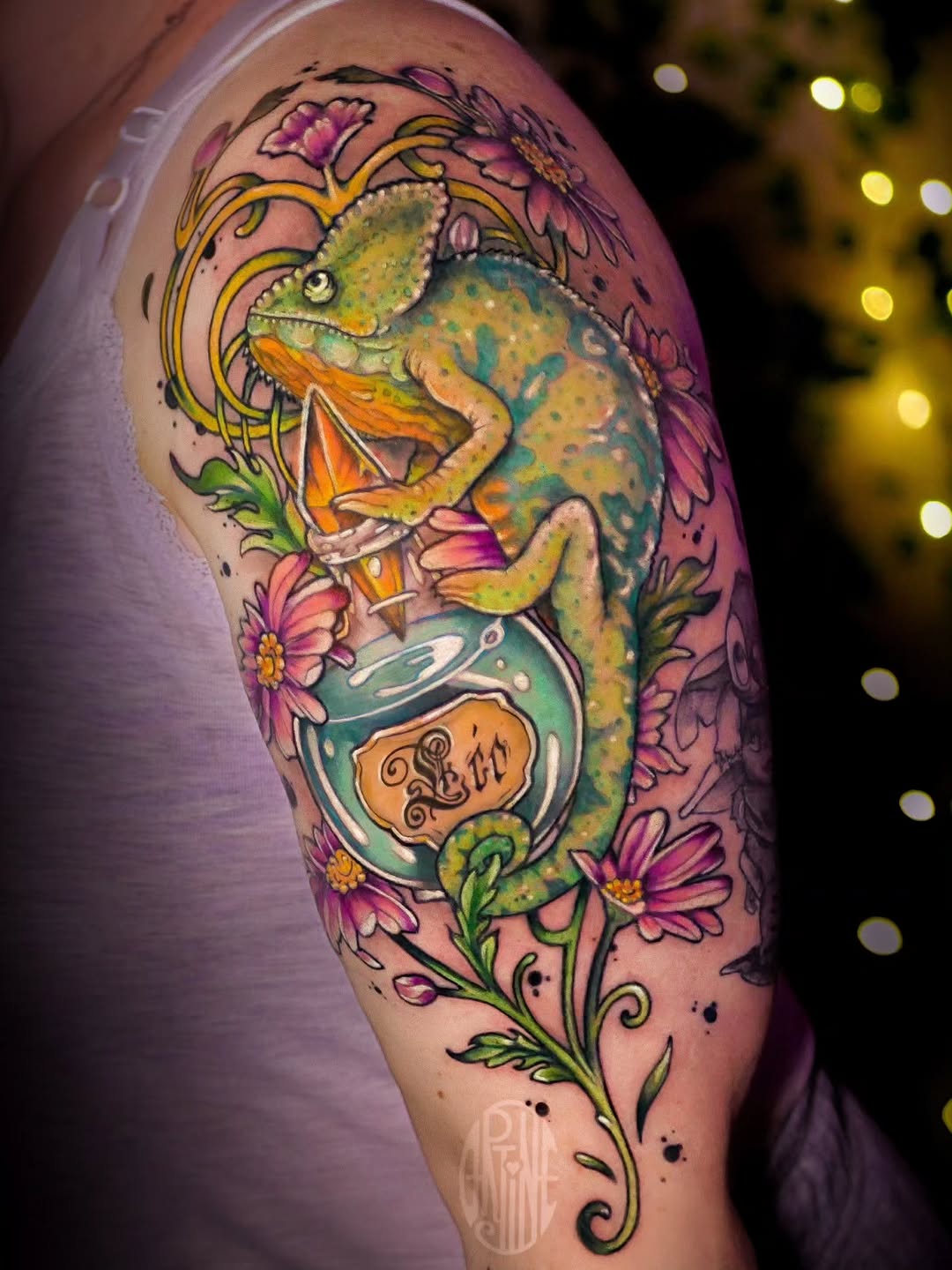 Colorful chameleon tattoo on upper arm holding a potion bottle, surrounded by pink and yellow flowers with green vines on pink skin.