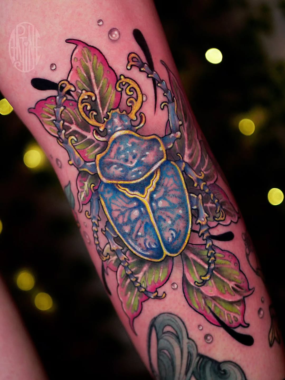 Colorful beetle tattoo with blue body, pink and magenta wings, gold ornamental details, and decorative flourishes on pink skin.