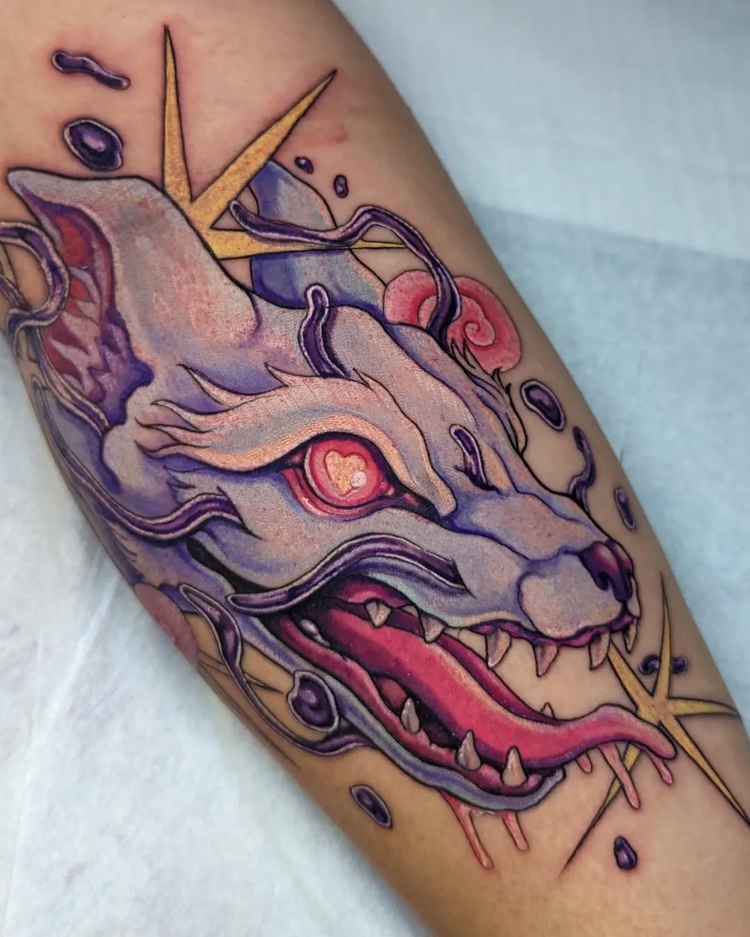 Colorful demon or dragon head tattoo on forearm featuring purple, red, and gold tones with sharp teeth, glowing eye, and decorative horns.