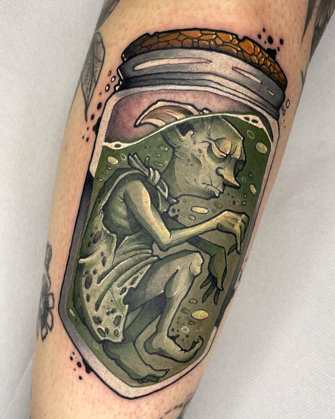 Colorful tattoo on an arm depicting a fairy or sprite figure seated inside an open glass jar with green liquid and floating particles, surrounded by a cork lid.