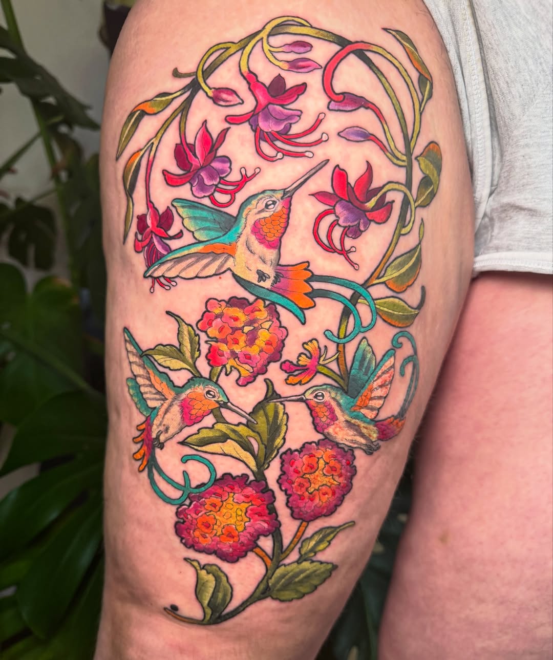 Colorful hummingbird tattoo on upper arm surrounded by pink and red flowers, green leaves, and curved vines in vibrant reds, oranges, teals, and golds.