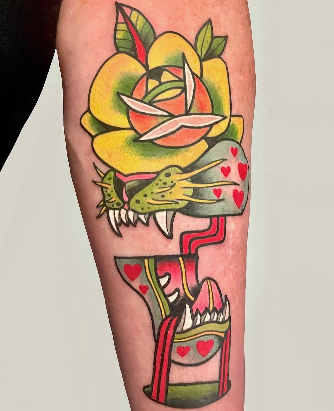 Colorful tattoo on a calf featuring a yellow rose with green leaves, a spotted bird with hearts, and a red container with hearts, outlined in black.