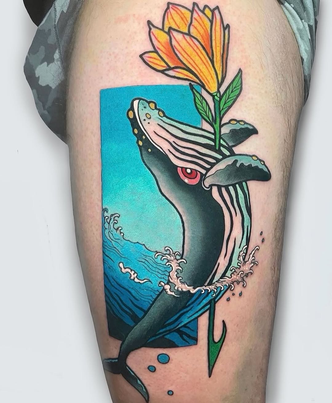 Whale breaching from turquoise water holds an orange flower in its mouth, tattooed on the thigh with vibrant colors and detailed linework.