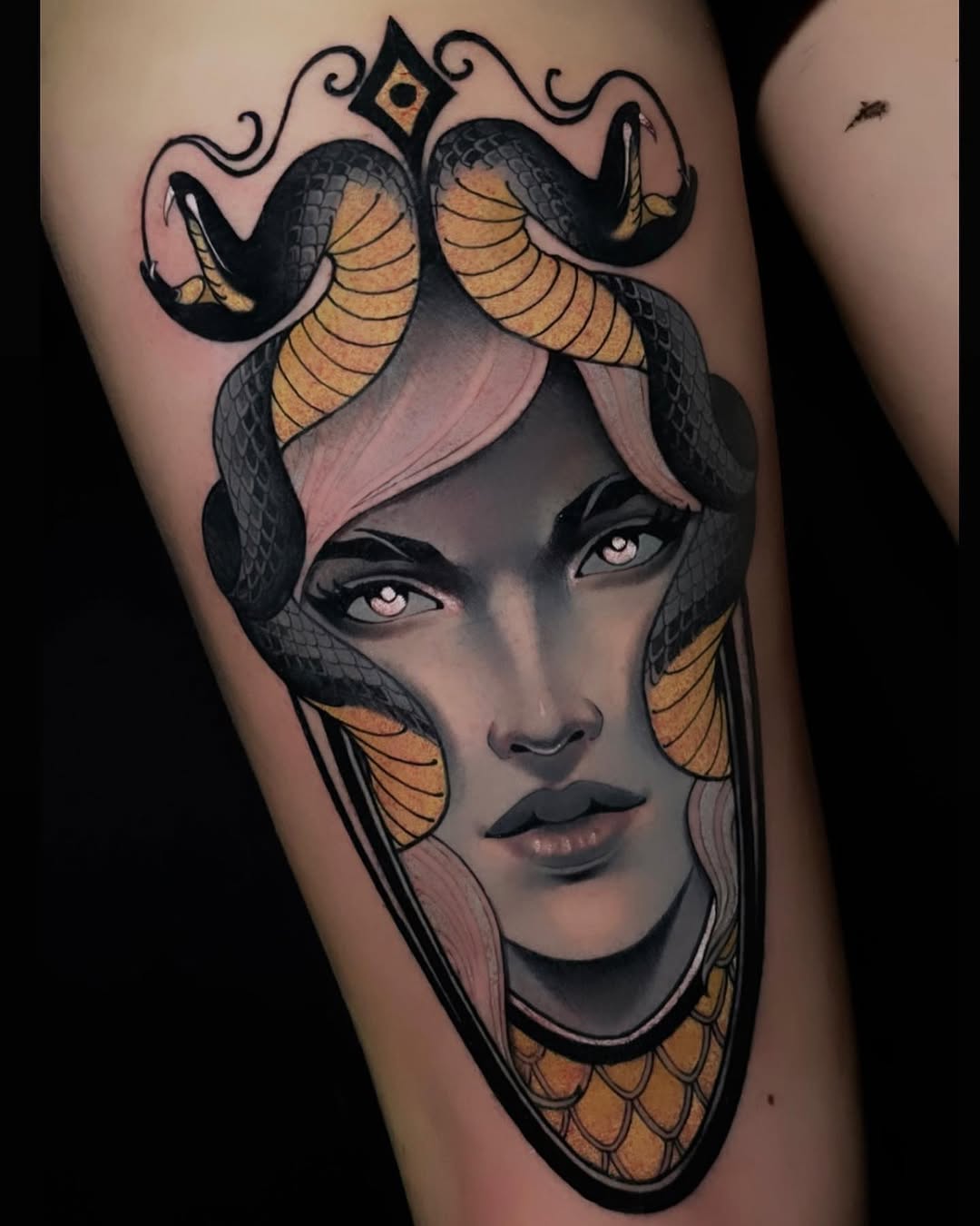 Medusa-inspired portrait tattoo on thigh featuring a woman's face with serpent hair in black, gold, and mauve tones, ornamental crown detail above.