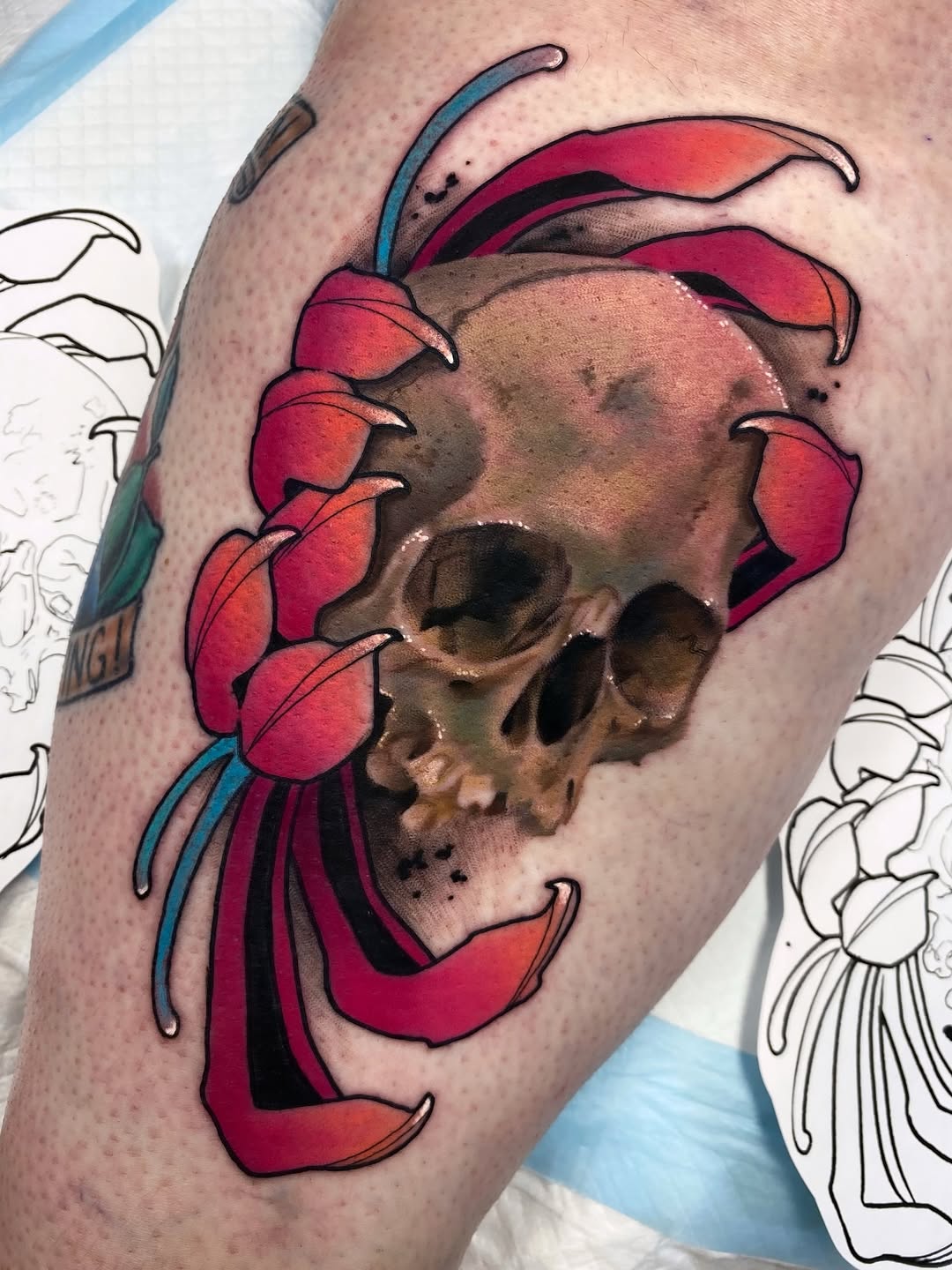 Colorful tattoo on upper arm featuring a skull with red flower petals and blue antennae surrounding it, displayed on light skin.
