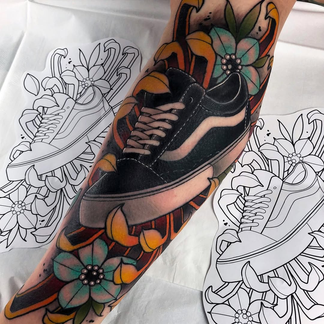 Colorful sleeve tattoo featuring a black sneaker surrounded by orange and teal flowers with decorative elements, displayed on an arm next to design sketches.