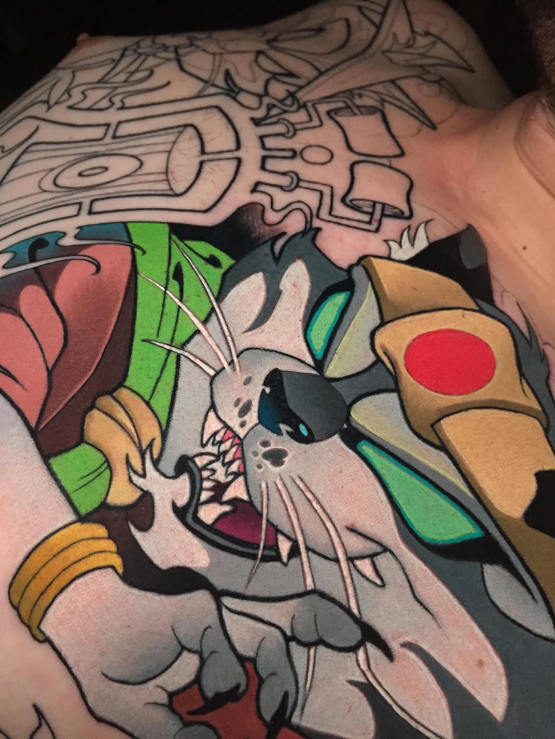 Colorful tattoo on shoulder featuring a rabbit character with green leaves, surrounded by abstract geometric line work in upper portion, predominantly green, red, tan, and black colors.