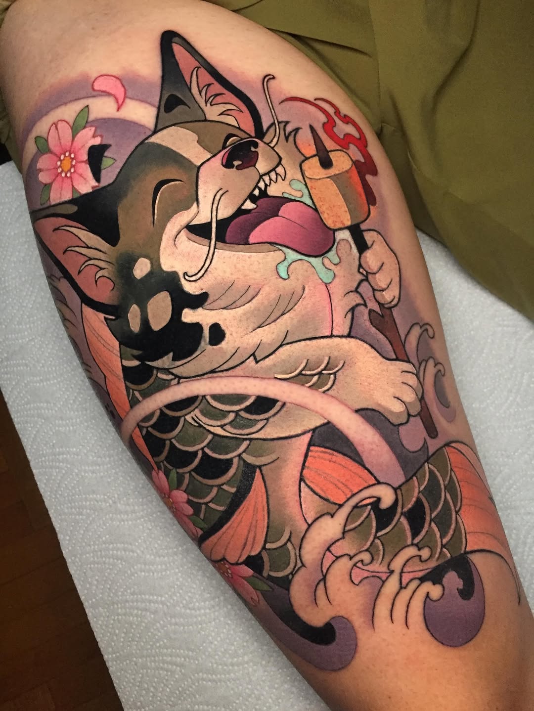 Colorful cat-fish hybrid tattoo on upper arm with open mouth, pink tongue, scales, flowers, and swirling water elements in orange, pink, and purple tones.