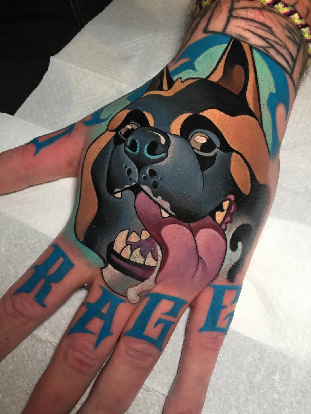 Colorful dog head tattoo on forearm and hand with blue text lettering, featuring brown and black tones with pink tongue and turquoise leaf details.