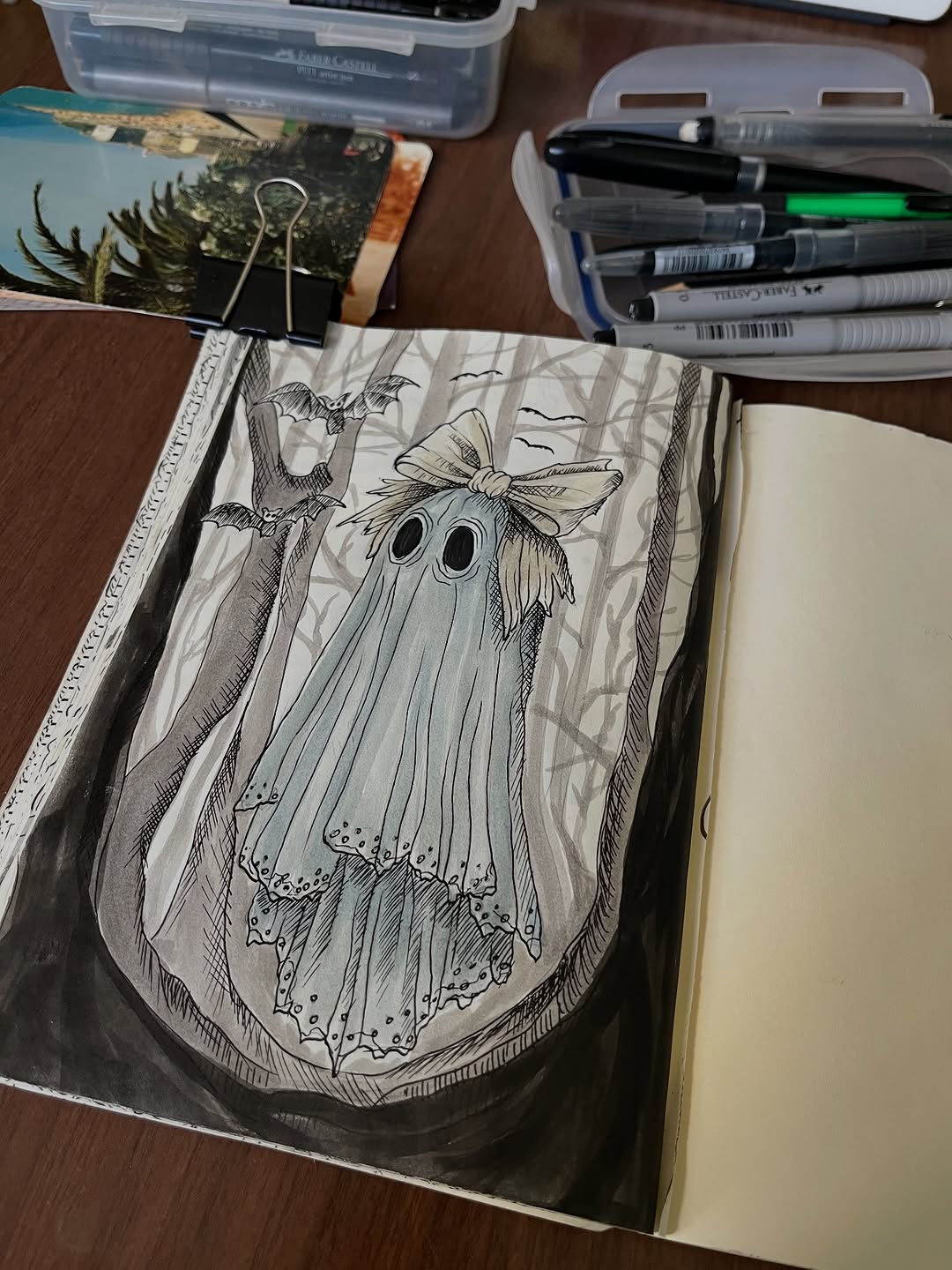 Sketch of a ghost with a bow and bats on lined paper, surrounded by markers and reference photos on a wooden desk.