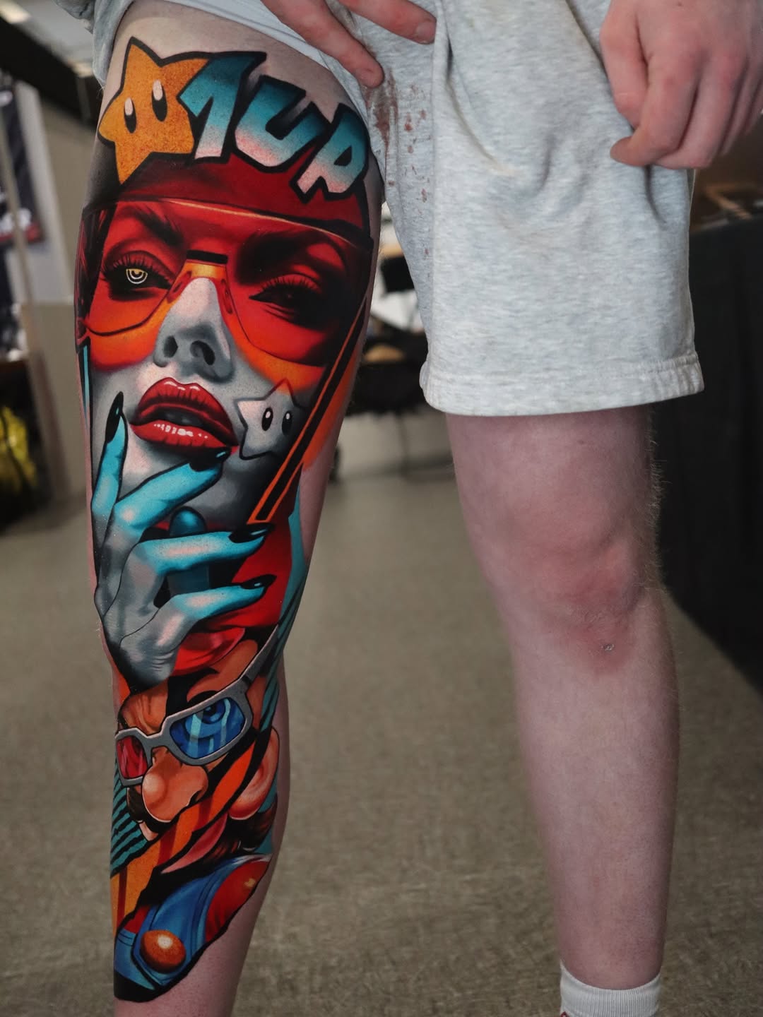 Full leg tattoo featuring a stylized female face with red glasses, blue hands, and geometric elements in red, blue, orange, and teal colors.