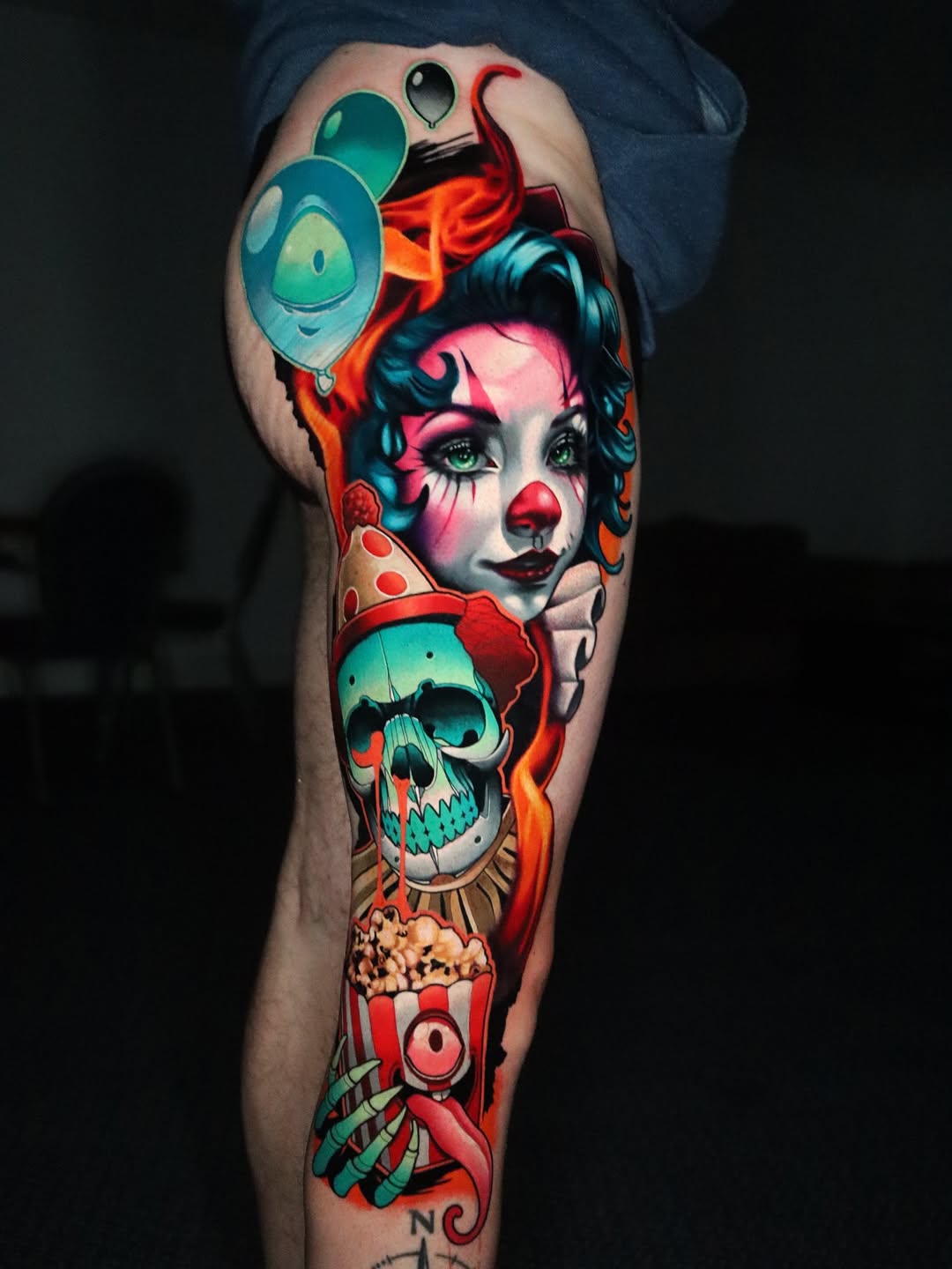 Colorful leg sleeve tattoo featuring a clown face, skull, balloons, popcorn, and circus elements in vibrant reds, teals, and oranges.