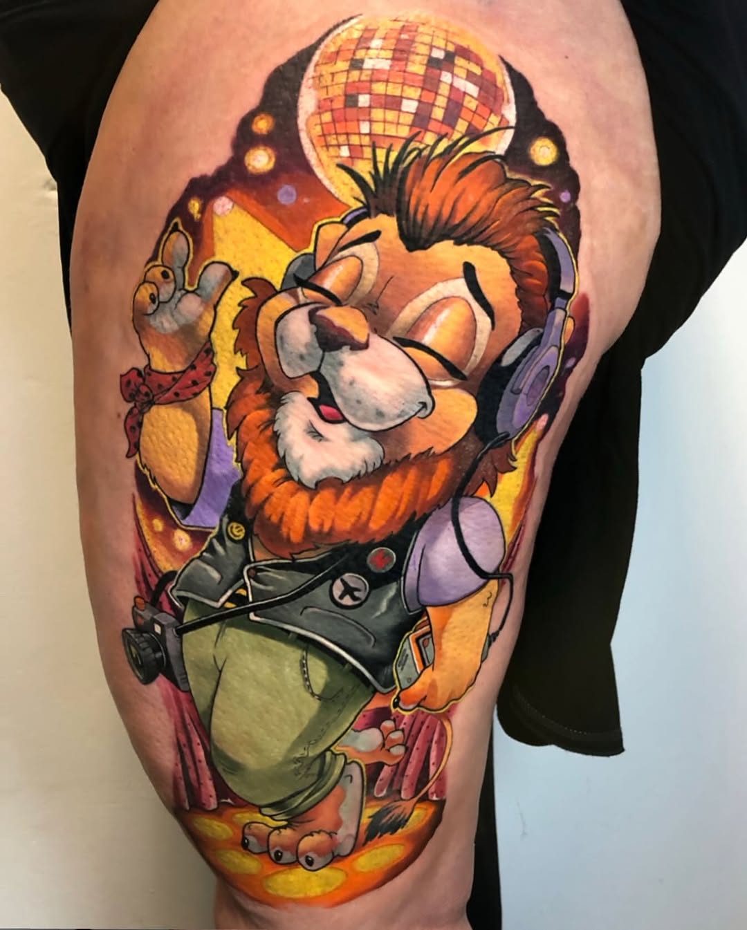 Colorful leg tattoo of an animated lion character wearing headphones and sunglasses, holding a disco ball, surrounded by warm orange, yellow, and purple tones.