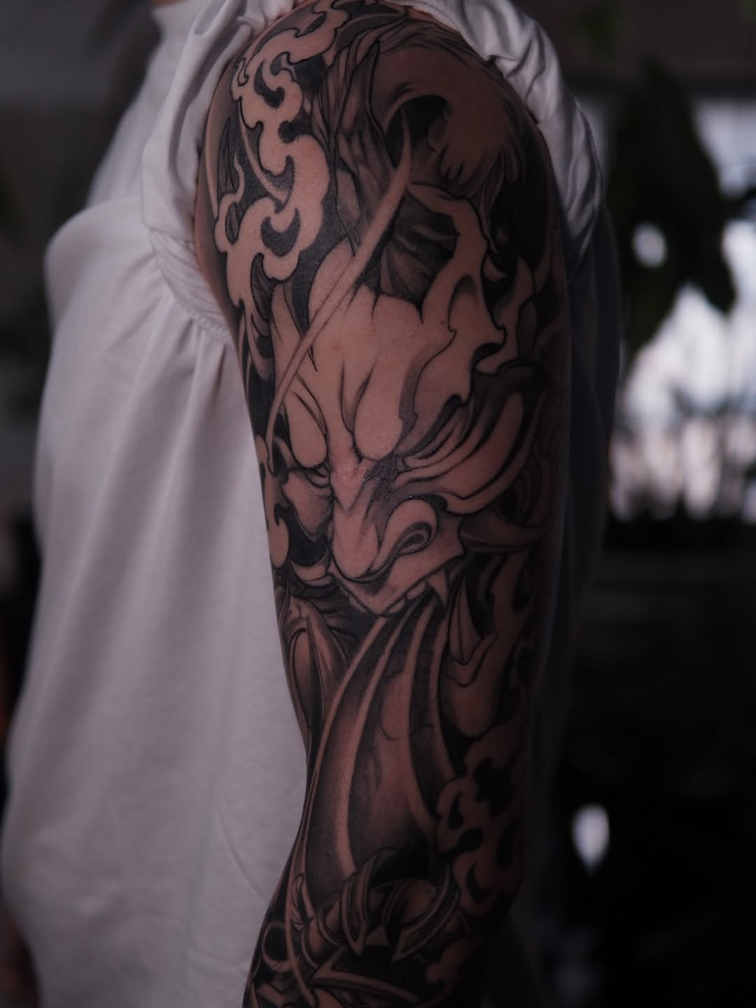 Detailed demon or oni mask tattoo on upper arm and shoulder with intricate line work and shading in dark ink on skin.