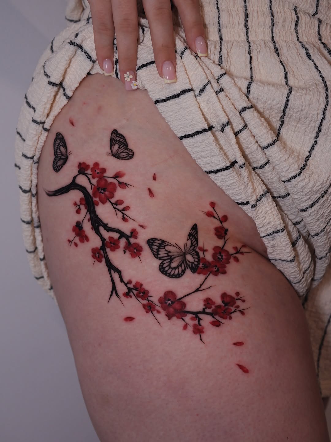 Thigh tattoo featuring black butterfly outlines and red cherry blossom branches with falling petals on skin.