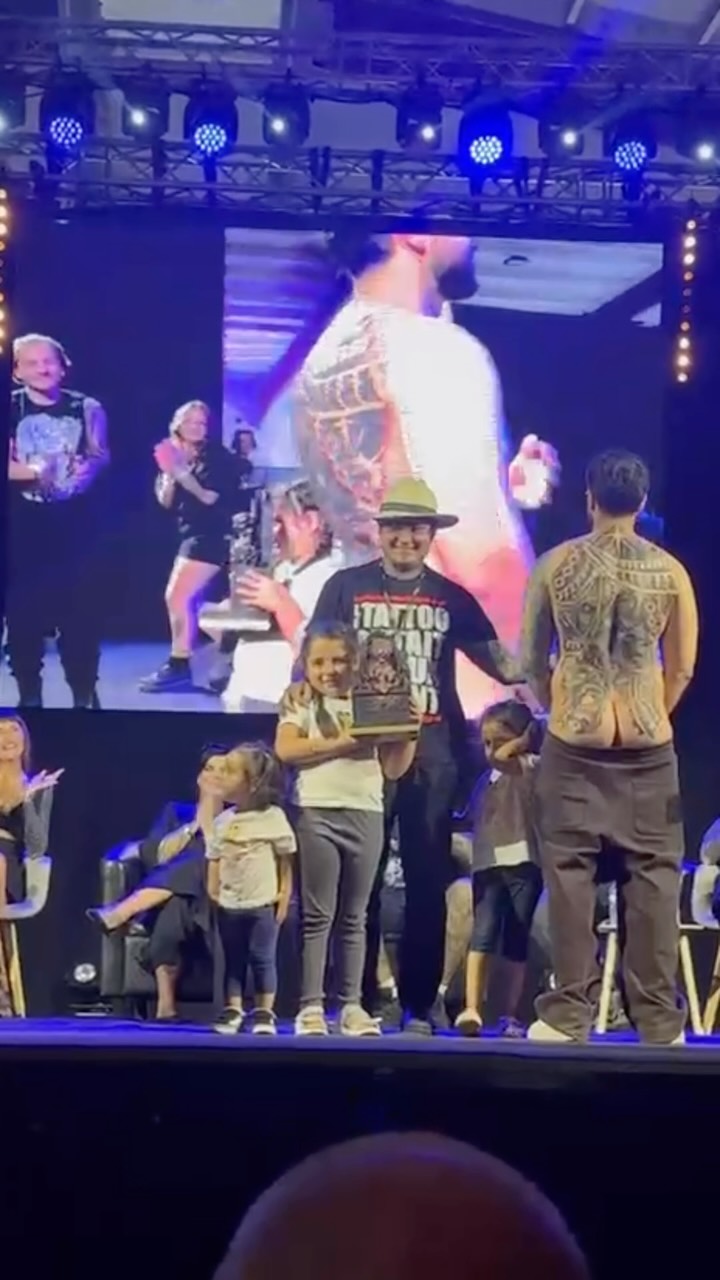Large-scale back tattoo featuring intricate golden and dark designs displayed on stage during a tattoo convention presentation with audience members present.