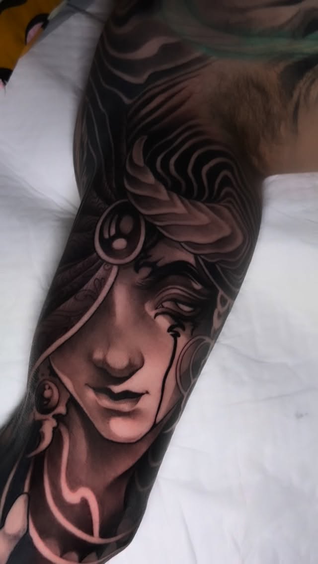 Detailed portrait tattoo on forearm featuring a classical female face with ornamental headdress and earrings in shades of brown and tan.