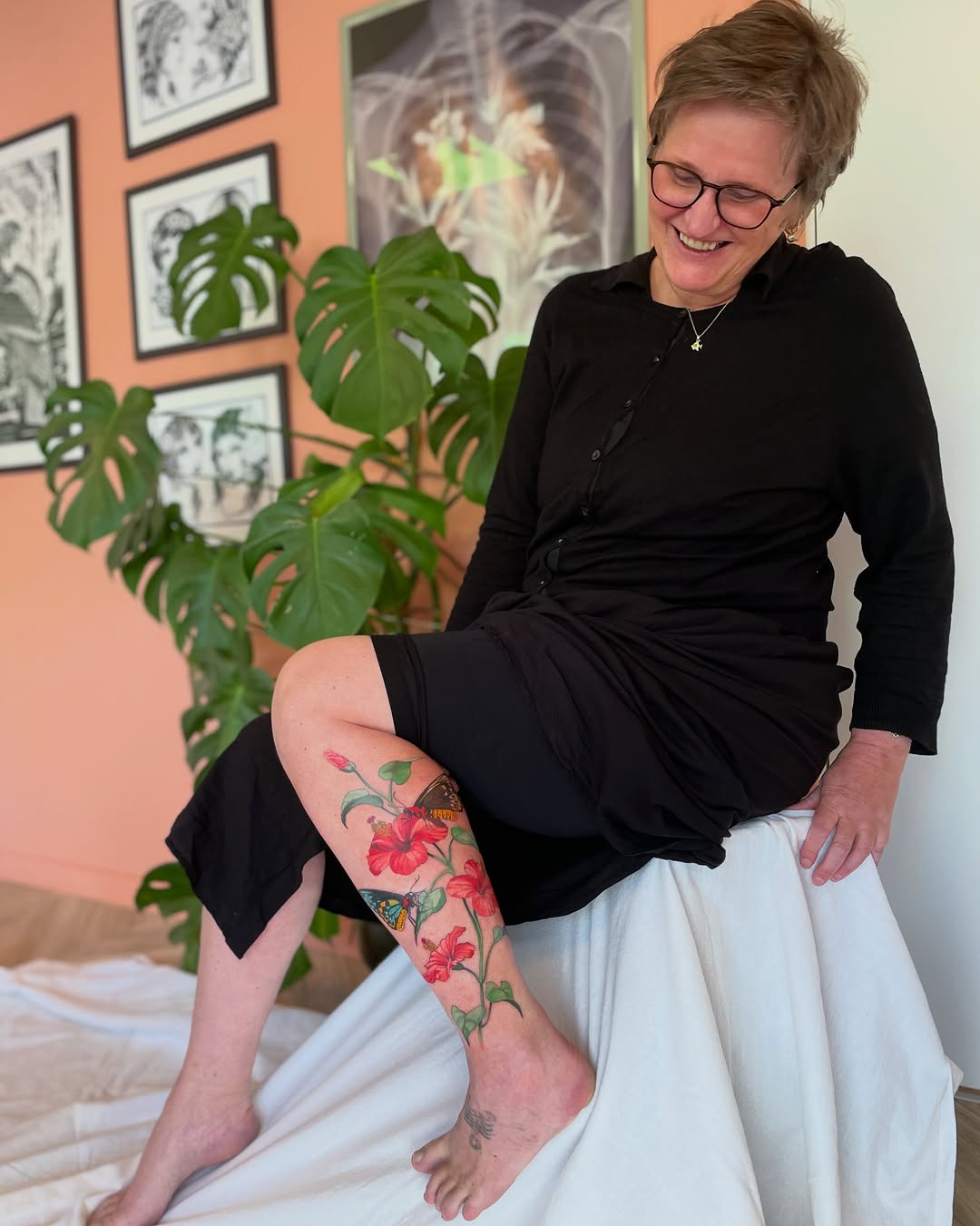 Woman displaying a colorful leg tattoo featuring red hibiscus flowers and green foliage wrapping around the calf and ankle area.