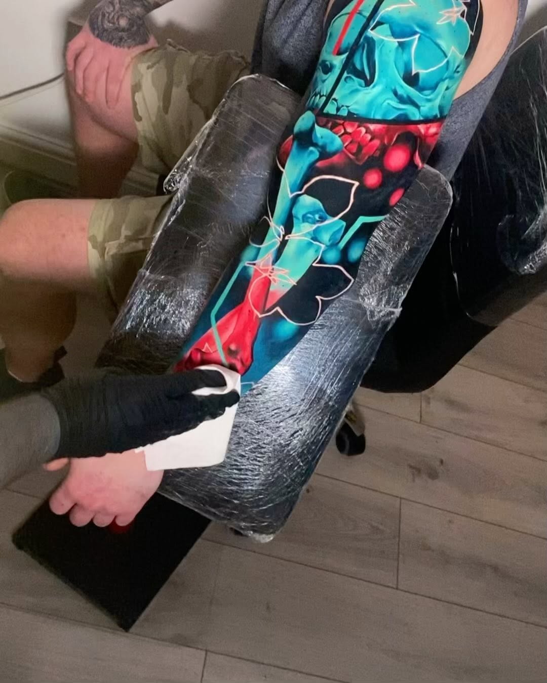 Colorful sleeve tattoo featuring blue and red floral designs with black outlines on the forearm, photographed during a consultation or fitting session.