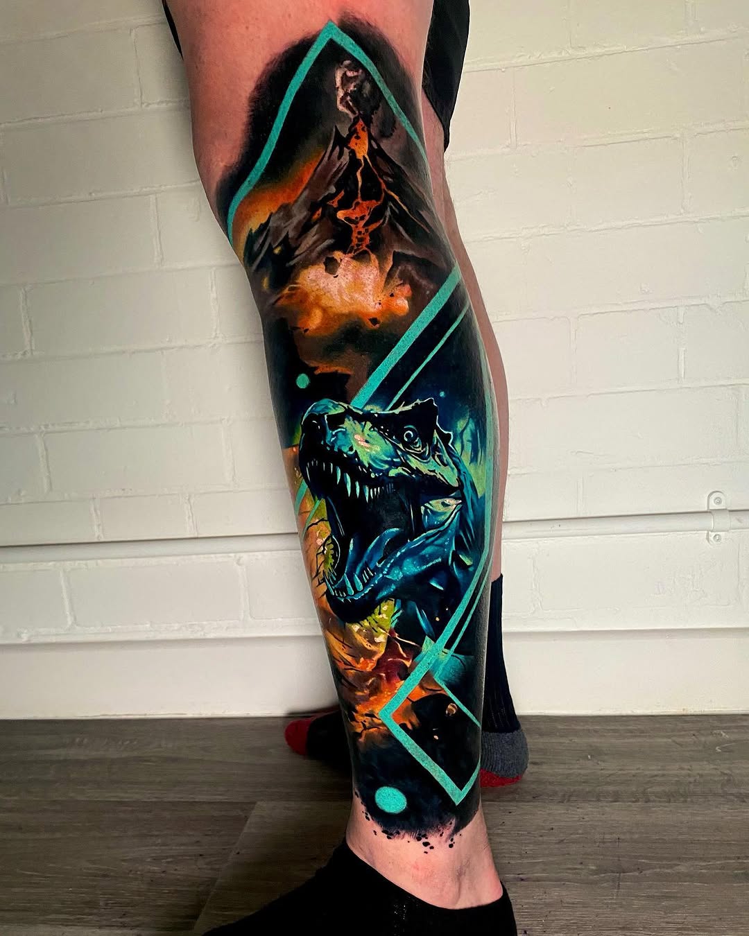 Colorful full-leg sleeve tattoo featuring a volcano, dinosaur head, and geometric turquoise shapes on dark background, displayed on calf and shin.