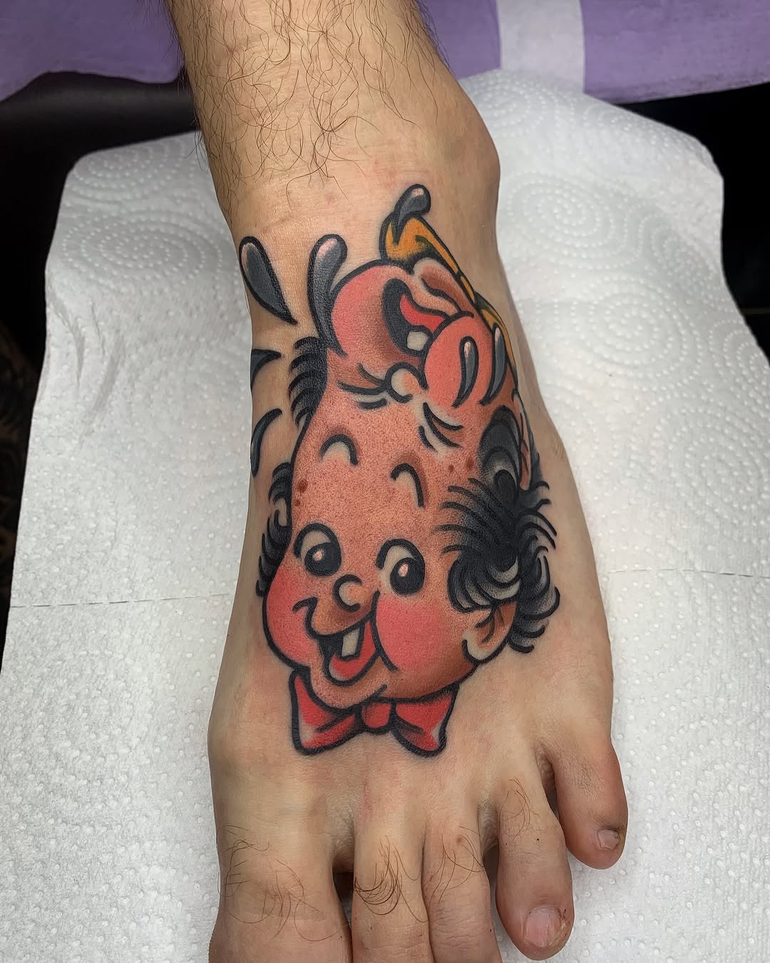 Colorful cartoon character head tattoo with red skin, black hair, bow tie, and smiling expression on the back of a hand.