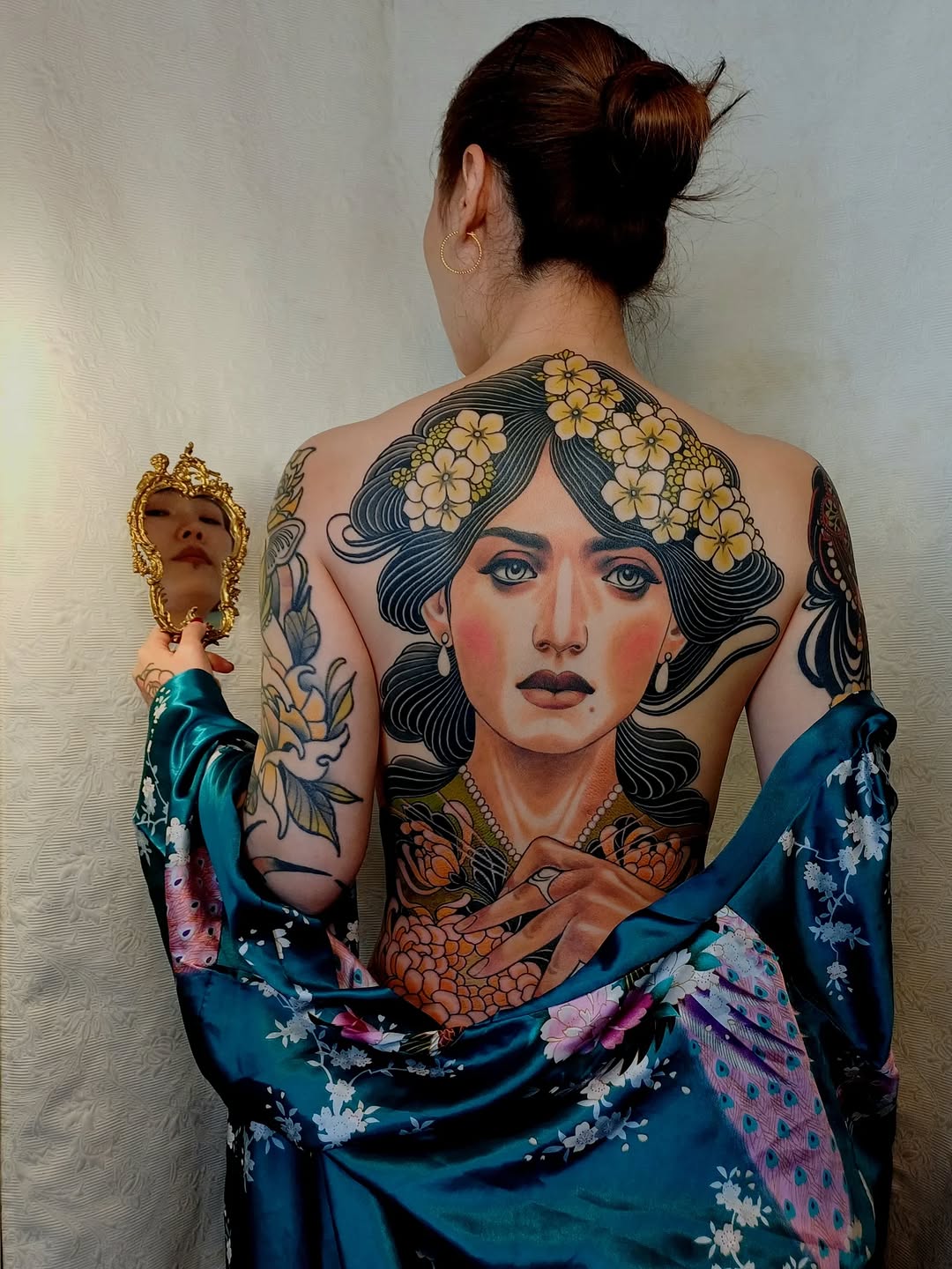 Woman displaying back and arm tattoos featuring a portrait of a woman with flowers in her hair and colored sleeve designs in blue, gold, and orange tones.