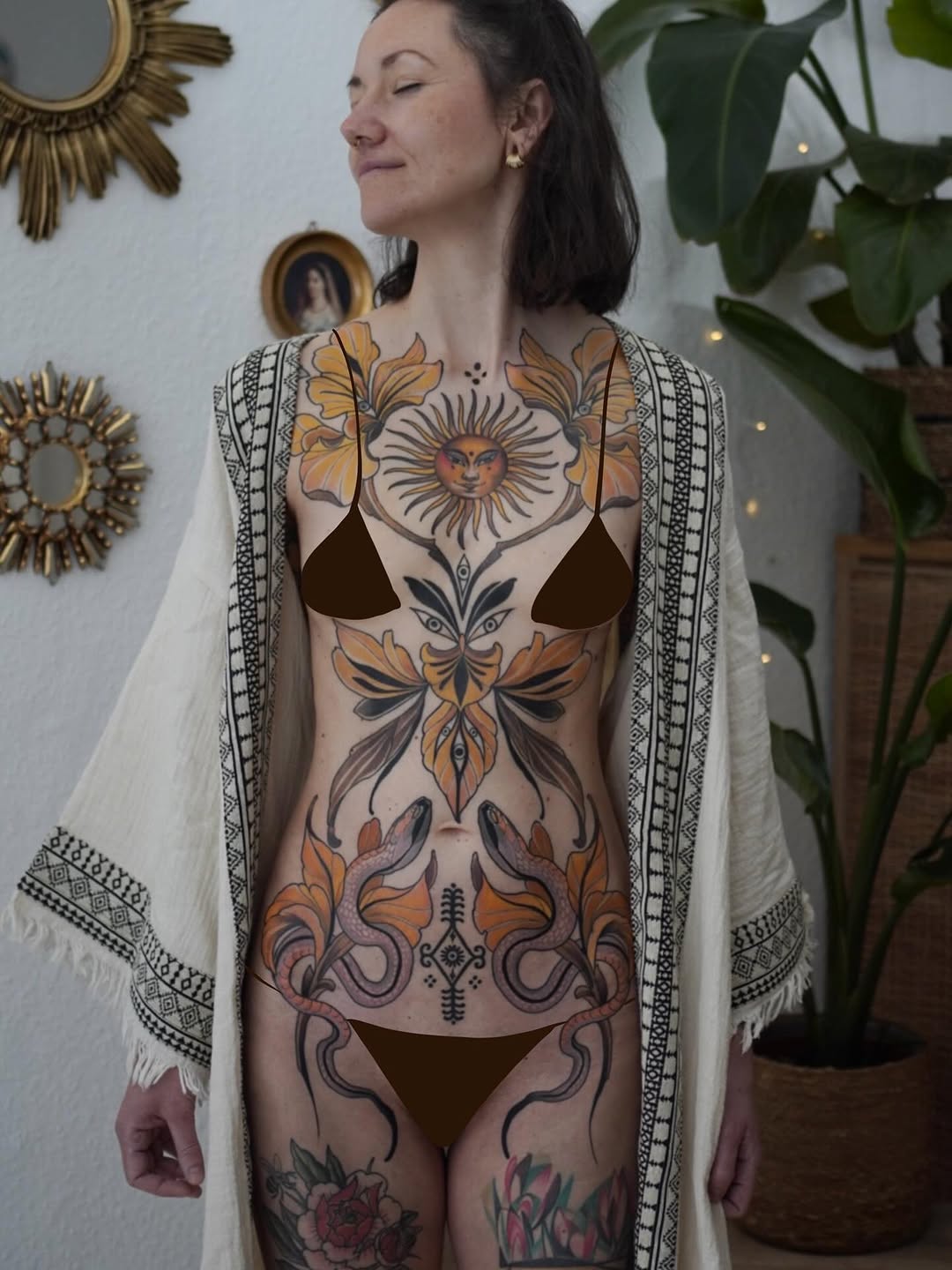 Woman displaying large-scale chest and torso tattoo featuring a celestial sun face, ornate floral motifs, and symmetrical designs in orange, black, and purple tones.