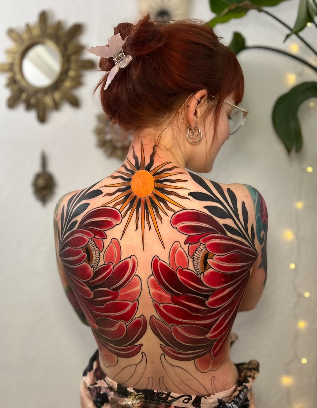 Back tattoo featuring symmetrical red flowers with an orange sun centerpiece and black botanical line work on a person with reddish-brown hair.