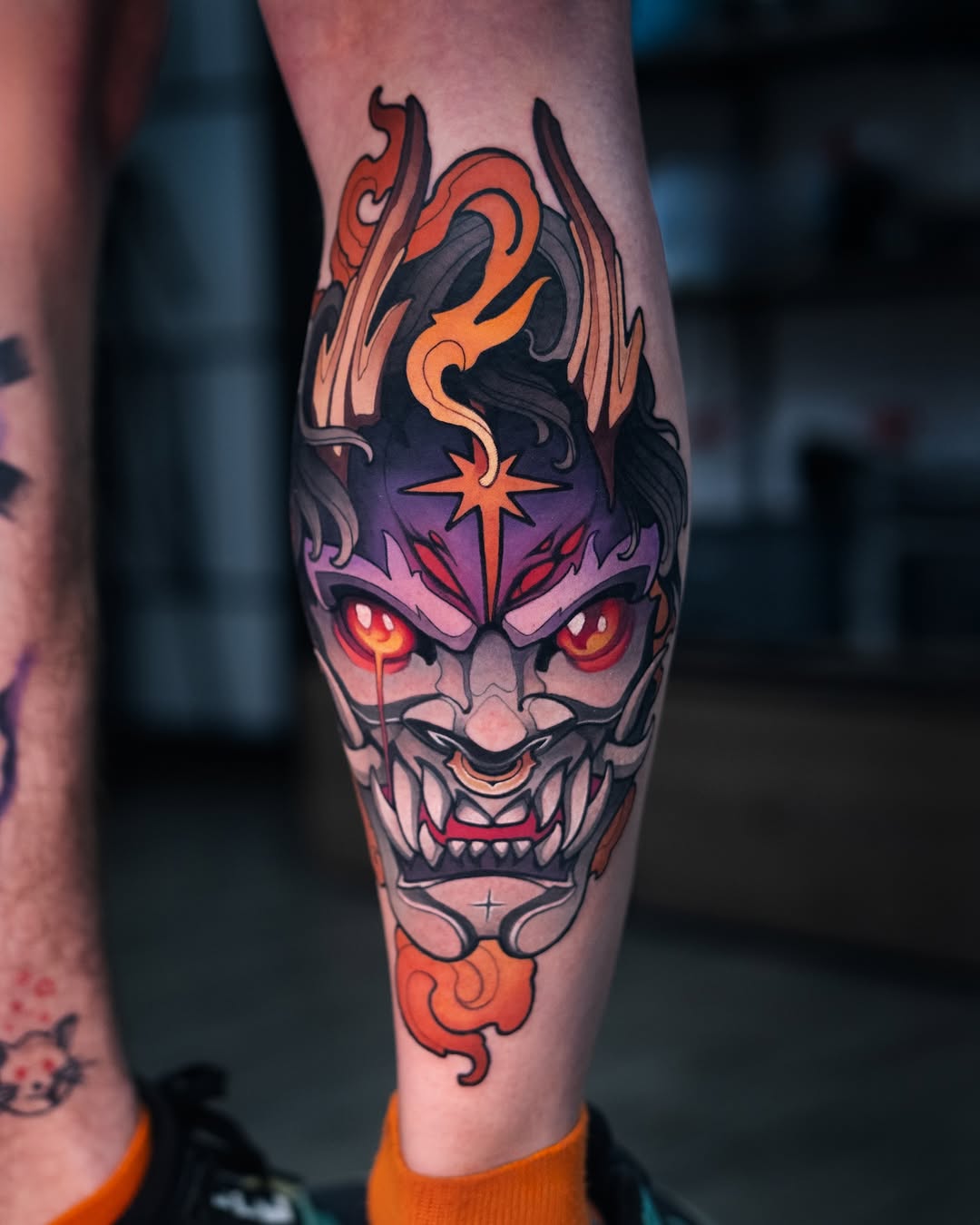 Demonic face tattoo with glowing red eyes, purple skin, orange flames, and a star marking on the forehead, placed on the calf.