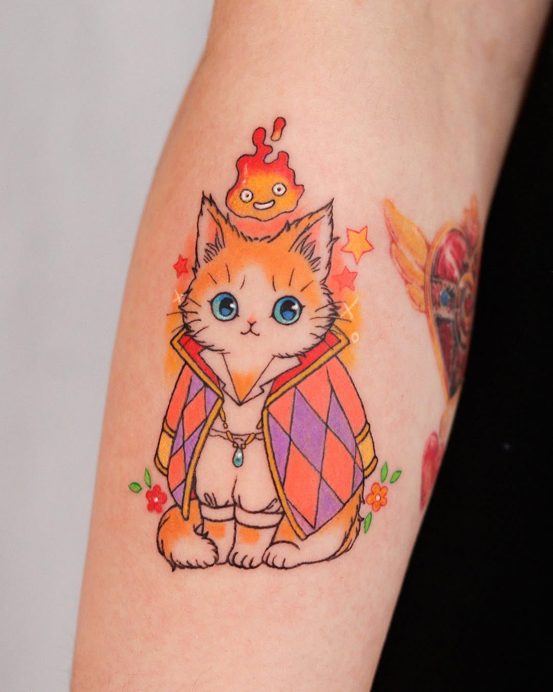 Orange tabby cat wearing a colorful diamond-patterned cape with a smiling flame character on its head, surrounded by stars and flowers, tattooed on an arm.